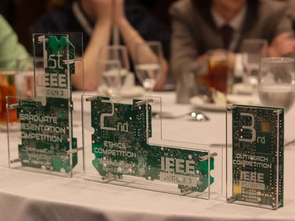 Circuit board plaques with 'IEEE' branding on a table with people in the background.